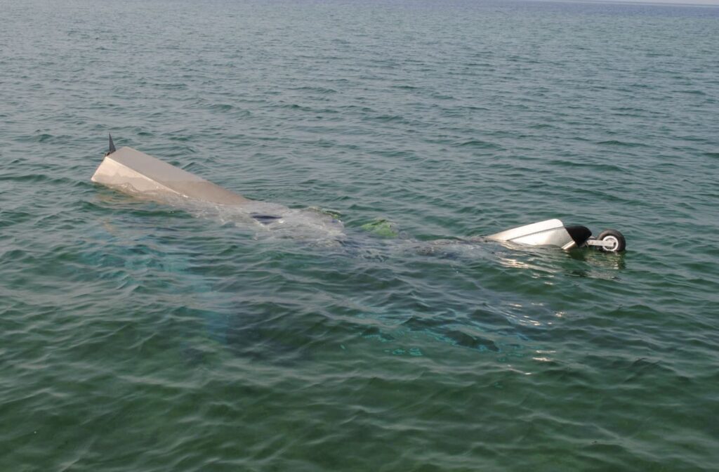 A portion of the aircraft wreckage, including a float and landing gear, partially submerged in the calm waters of Lake Michigan. The surface is glassy, and the horizon is faint in the distance under hazy conditions.